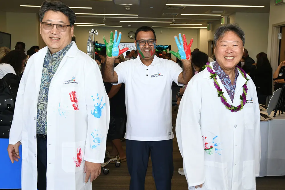 Three smiling men two doctors with colorful handprints on their white coats and one in the center holding up multicolor painted hands. 
