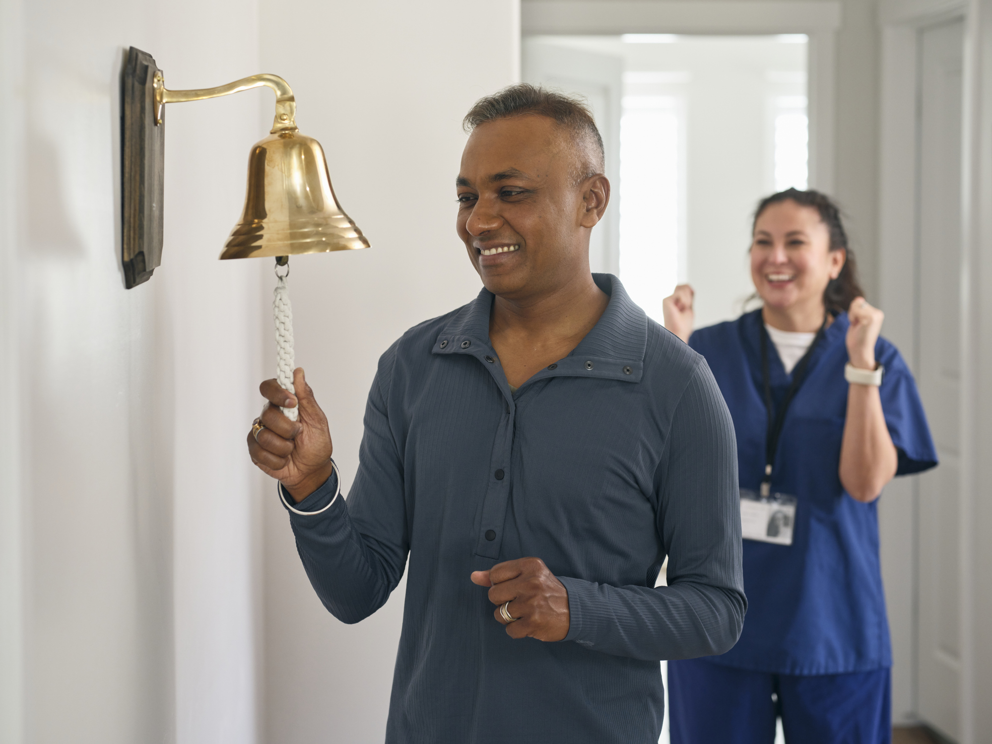 An man rings the bell in a treatment office to signify the end of his chemotherapy treatment office, as his patient navigator cheers in celebration