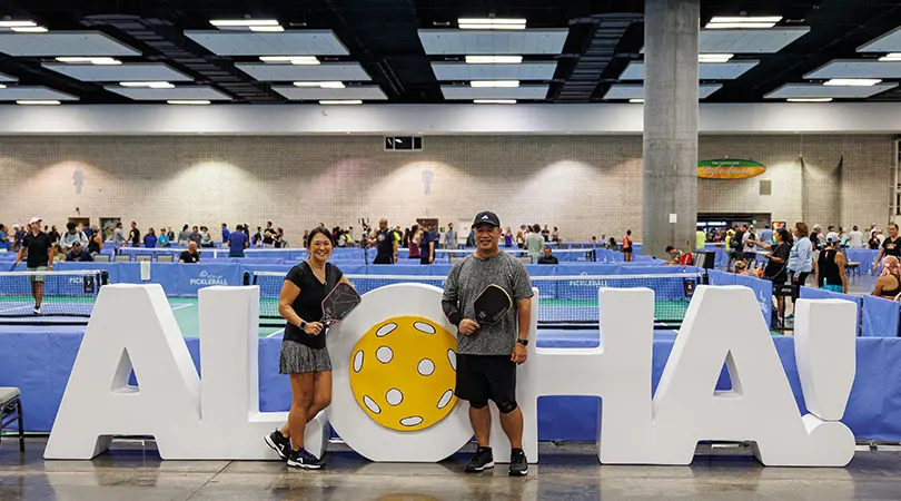 Man and woman holding pickleball paddles in front of large ALOHA letters with pickleball courts in the background.