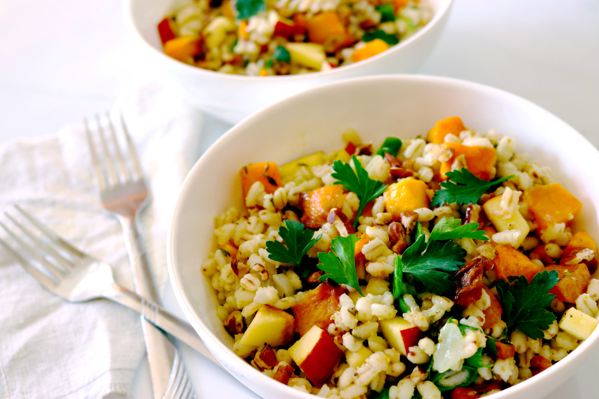 Two white bowls filled with Fall Farro Salad sitting on a white marble countertop with two forks and a burlap napkin placed next to the bowls.